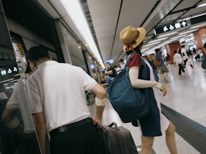 People in a bustling subway or train station, with one individual wearing a straw hat and a blue backpack. Another person is holding a suitcase near the platform edge. Signs and lights are visible, reflecting the busy transport environment.