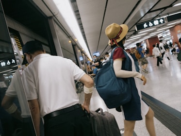 People in a bustling subway or train station, with one individual wearing a straw hat and a blue backpack. Another person is holding a suitcase near the platform edge. Signs and lights are visible, reflecting the busy transport environment.