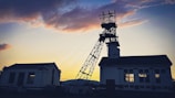 A silhouetted industrial structure stands against a dramatic sunset sky, with hues of orange and purple. The building appears to be a mining facility with a tall headframe, and there is also a secondary building with a pitched roof visible.