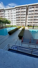 A modern apartment building with balconies overlooking a swimming pool area. The area has two swimming pools separated by a walkway lined with trimmed hedges. Traffic cones are placed near the walkway for safety. The sky is clear with few clouds.