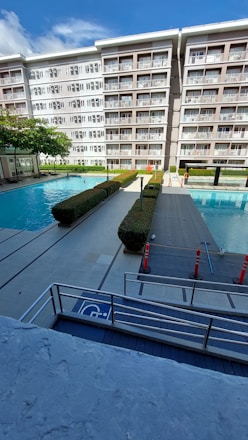 A modern apartment building with balconies overlooking a swimming pool area. The area has two swimming pools separated by a walkway lined with trimmed hedges. Traffic cones are placed near the walkway for safety. The sky is clear with few clouds.
