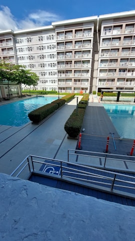 A modern apartment building with balconies overlooking a swimming pool area. The area has two swimming pools separated by a walkway lined with trimmed hedges. Traffic cones are placed near the walkway for safety. The sky is clear with few clouds.