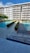 A modern apartment building with balconies overlooking a swimming pool area. The area has two swimming pools separated by a walkway lined with trimmed hedges. Traffic cones are placed near the walkway for safety. The sky is clear with few clouds.