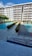 A modern apartment building with balconies overlooking a swimming pool area. The area has two swimming pools separated by a walkway lined with trimmed hedges. Traffic cones are placed near the walkway for safety. The sky is clear with few clouds.