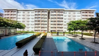 A modern residential building with six floors and numerous balconies is prominently visible against a blue sky. In front of the building, there are two large outdoor swimming pools surrounded by neatly arranged pathways and hedges. Several trees and manicured bushes are scattered throughout the area, and a few people can be seen walking along the pathways or swimming.