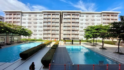 A modern residential building with six floors and numerous balconies is prominently visible against a blue sky. In front of the building, there are two large outdoor swimming pools surrounded by neatly arranged pathways and hedges. Several trees and manicured bushes are scattered throughout the area, and a few people can be seen walking along the pathways or swimming.