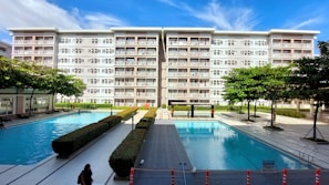A modern residential building with six floors and numerous balconies is prominently visible against a blue sky. In front of the building, there are two large outdoor swimming pools surrounded by neatly arranged pathways and hedges. Several trees and manicured bushes are scattered throughout the area, and a few people can be seen walking along the pathways or swimming.