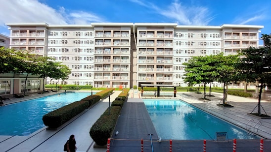 A modern residential building with six floors and numerous balconies is prominently visible against a blue sky. In front of the building, there are two large outdoor swimming pools surrounded by neatly arranged pathways and hedges. Several trees and manicured bushes are scattered throughout the area, and a few people can be seen walking along the pathways or swimming.