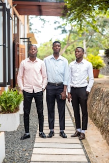 Three young men are standing on a walkway surrounded by greenery. They are dressed in smart casual attire, including neatly pressed shirts and trousers. The backdrop features a modern building with glass doors and potted plants.