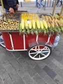 A street vendor cart with a variety of cooked and raw corn, as well as roasted chestnuts. The cart is red with white accents and has large wheels. A person is serving the food, with a road and pedestrians visible in the background.