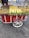 A street vendor cart with a variety of cooked and raw corn, as well as roasted chestnuts. The cart is red with white accents and has large wheels. A person is serving the food, with a road and pedestrians visible in the background.
