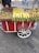 A street vendor cart with a variety of cooked and raw corn, as well as roasted chestnuts. The cart is red with white accents and has large wheels. A person is serving the food, with a road and pedestrians visible in the background.