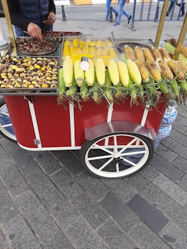 A street vendor cart with a variety of cooked and raw corn, as well as roasted chestnuts. The cart is red with white accents and has large wheels. A person is serving the food, with a road and pedestrians visible in the background.