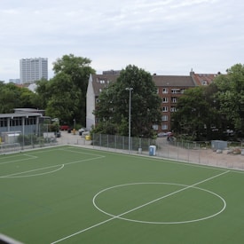 A sports field with a green artificial turf is surrounded by a metal fence. The area is empty with no people visible. In the background, there are several residential buildings with multi-story apartments, characterized by brick and concrete architecture. Tall trees are present around the buildings, providing greenery to the urban setting. A cloudy sky looms overhead.