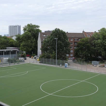 A sports field with a green artificial turf is surrounded by a metal fence. The area is empty with no people visible. In the background, there are several residential buildings with multi-story apartments, characterized by brick and concrete architecture. Tall trees are present around the buildings, providing greenery to the urban setting. A cloudy sky looms overhead.