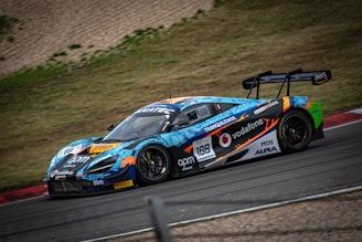 A vibrant race car, adorned with colorful graphics and sponsor logos, speeds around a racetrack. The vehicle features a prominent rear spoiler and sleek design, optimized for high performance. Surrounding the track is a grassy area and gravel.