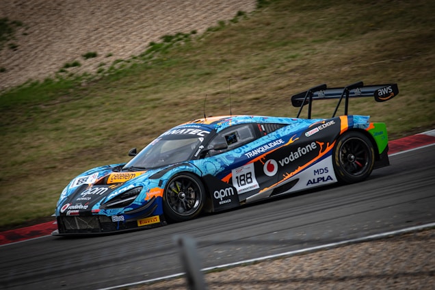 A vibrant race car, adorned with colorful graphics and sponsor logos, speeds around a racetrack. The vehicle features a prominent rear spoiler and sleek design, optimized for high performance. Surrounding the track is a grassy area and gravel.