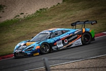 A vibrant race car, adorned with colorful graphics and sponsor logos, speeds around a racetrack. The vehicle features a prominent rear spoiler and sleek design, optimized for high performance. Surrounding the track is a grassy area and gravel.