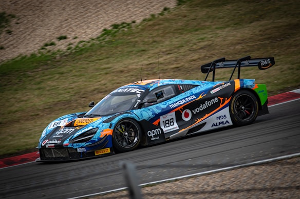 A vibrant race car, adorned with colorful graphics and sponsor logos, speeds around a racetrack. The vehicle features a prominent rear spoiler and sleek design, optimized for high performance. Surrounding the track is a grassy area and gravel.