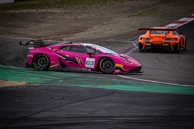 Two racing cars are captured on a track, one prominently pink and the other orange. The pink car, with the number 83, is sharply detailed in the foreground, showcasing sponsorship decals and aerodynamic design. The track features both asphalt and green curbing, indicating a race setting.
