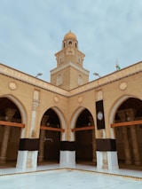 A tan brick mosque with an ornate dome rises above a courtyard. Arched entrances are adorned with intricate designs, and a decorative black cloth is draped over pillars. The structure is accented with detailed carvings and Arabic script.