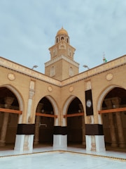 A tan brick mosque with an ornate dome rises above a courtyard. Arched entrances are adorned with intricate designs, and a decorative black cloth is draped over pillars. The structure is accented with detailed carvings and Arabic script.
