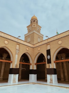 A tan brick mosque with an ornate dome rises above a courtyard. Arched entrances are adorned with intricate designs, and a decorative black cloth is draped over pillars. The structure is accented with detailed carvings and Arabic script.