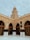 A tan brick mosque with an ornate dome rises above a courtyard. Arched entrances are adorned with intricate designs, and a decorative black cloth is draped over pillars. The structure is accented with detailed carvings and Arabic script.