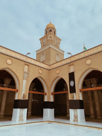 A tan brick mosque with an ornate dome rises above a courtyard. Arched entrances are adorned with intricate designs, and a decorative black cloth is draped over pillars. The structure is accented with detailed carvings and Arabic script.
