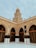 A tan brick mosque with an ornate dome rises above a courtyard. Arched entrances are adorned with intricate designs, and a decorative black cloth is draped over pillars. The structure is accented with detailed carvings and Arabic script.