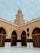 A tan brick mosque with an ornate dome rises above a courtyard. Arched entrances are adorned with intricate designs, and a decorative black cloth is draped over pillars. The structure is accented with detailed carvings and Arabic script.