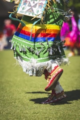 A person is wearing an ornate and colorful traditional outfit with a green fabric base and vibrant horizontal stripes in red, orange, yellow, and blue. The outfit is embellished with silver cone decorations and fringes at the hem. The person is in motion, wearing red moccasins while performing a dance on a grassy field.