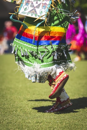 A person is wearing an ornate and colorful traditional outfit with a green fabric base and vibrant horizontal stripes in red, orange, yellow, and blue. The outfit is embellished with silver cone decorations and fringes at the hem. The person is in motion, wearing red moccasins while performing a dance on a grassy field.