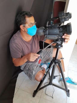 A person wearing a blue face mask and glasses is squatting beside a professional video camera mounted on a tripod. The person is casually dressed in a brown t-shirt and gray shorts, focusing intently on the camera setup. The background is simple and unadorned, with a dark cloth backdrop.