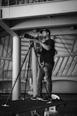 A man stands on a platform, holding a camera with a large telephoto lens mounted on a tripod. He wears a cap, glasses, and casual attire, appearing focused on photographing something off-frame. The setting is indoors, with industrial architecture, including metal beams and fabric canopy structures in the background.