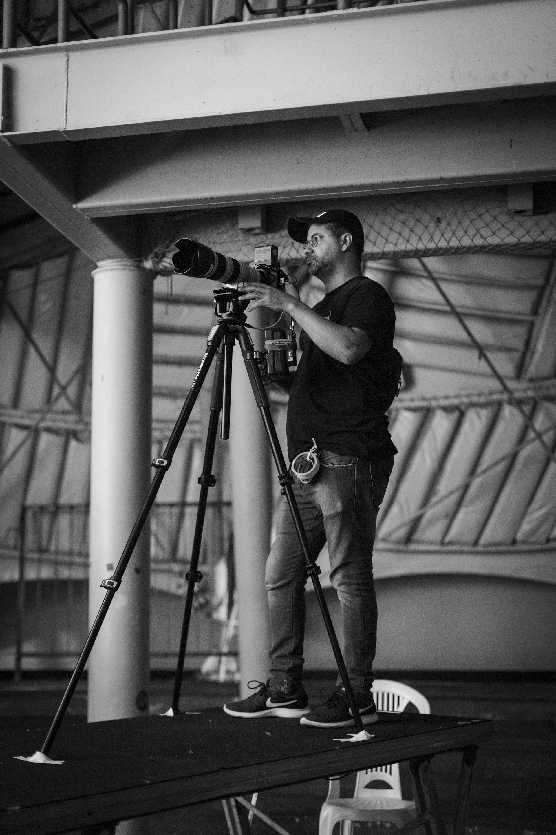 A man stands on a platform, holding a camera with a large telephoto lens mounted on a tripod. He wears a cap, glasses, and casual attire, appearing focused on photographing something off-frame. The setting is indoors, with industrial architecture, including metal beams and fabric canopy structures in the background.