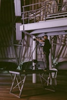 A person stands on a wooden platform, operating a camera mounted on a tripod in an industrial or warehouse-like setting. The structure features exposed beams and metal railings, while the floor appears to be wooden with a plastic chair nearby.