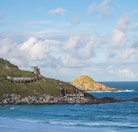 A coastal scene featuring a collection of small, colorful houses or huts lining the rocky shoreline. In the background, an old, abandoned building structure sits atop a grassy hill, with another rocky hill extending into the ocean. The ocean waves calmly crash against the shore under a partly cloudy sky.