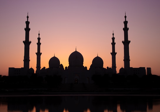 A silhouetted view of a grand mosque against a vibrant sunset sky. The structure features multiple minarets and domes, creating an impressive architectural silhouette. The sky transitions from warm orange to deep purple, casting a serene and majestic aura around the building.