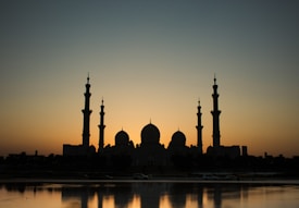 Silhouette of a grand mosque with multiple domes and tall minarets against the backdrop of a setting or rising sun, creating a striking contrast in the sky. The mosque is reflected on a body of water in the foreground, adding to the serene and peaceful atmosphere.
