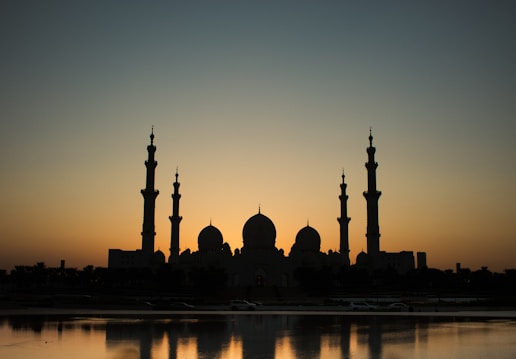 Silhouette of a grand mosque with multiple domes and tall minarets against the backdrop of a setting or rising sun, creating a striking contrast in the sky. The mosque is reflected on a body of water in the foreground, adding to the serene and peaceful atmosphere.