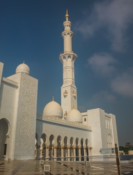 The image displays a grand mosque with intricate architectural features, including a tall minaret, white domes, and detailed carvings on the walls. The structure is set against a clear blue sky, giving it an elegant and majestic appearance.
