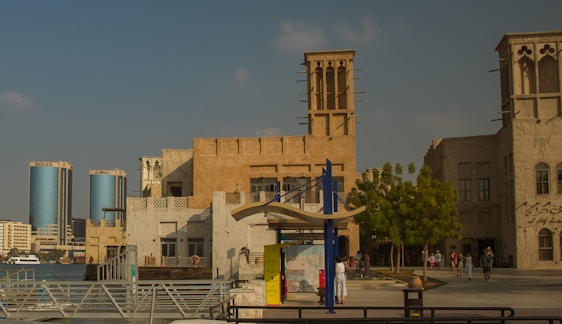 An urban scene with traditional Middle Eastern architecture in the foreground, including beige buildings with wind towers. A modern cityscape with tall, cylindrical skyscrapers can be seen in the background. The setting appears to be by the waterfront with trees, people walking, and a few boats on the water.