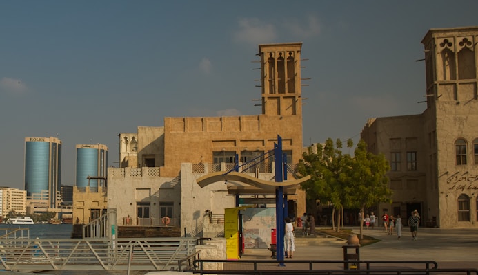 An urban scene with traditional Middle Eastern architecture in the foreground, including beige buildings with wind towers. A modern cityscape with tall, cylindrical skyscrapers can be seen in the background. The setting appears to be by the waterfront with trees, people walking, and a few boats on the water.