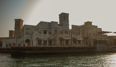 A traditional architectural building with ornate details and wind towers is situated by the water. The structure is composed of sand-colored stone and features several arched windows and doors. There are small signs of shops or cafes on the ground level. The setting sun casts a warm glow over the scene, creating long shadows and illuminating the intricate design of the facade.