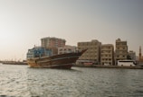 A traditional wooden dhow is floating on the water near the shoreline of a city. The cityscape includes a series of mid-rise buildings with a modern architectural style. A mosque with a minaret is visible in the background, further enriching the cultural atmosphere. The scene is set against a clear sky, and the water appears calm.