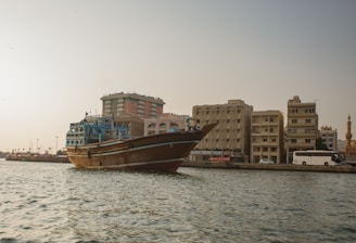 A traditional wooden dhow is floating on the water near the shoreline of a city. The cityscape includes a series of mid-rise buildings with a modern architectural style. A mosque with a minaret is visible in the background, further enriching the cultural atmosphere. The scene is set against a clear sky, and the water appears calm.