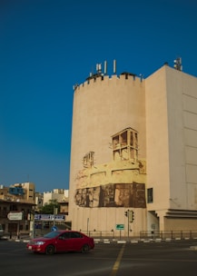 A large building features a cylindrical section with a mural depicting abstract architectural elements. The scene includes a red car driving on the street below, with additional buildings and street signs in the background under a clear blue sky.