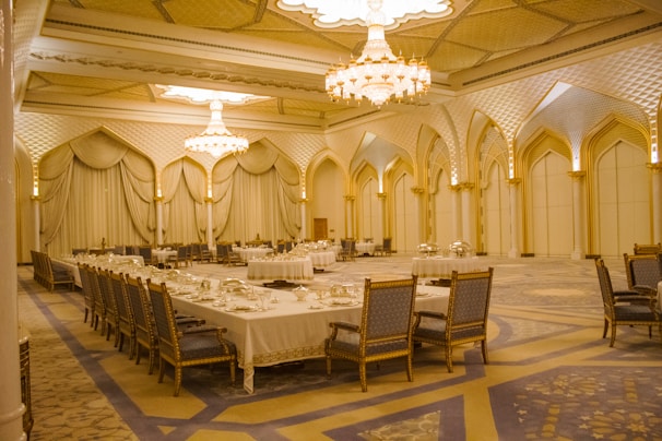 An opulent banquet hall adorned with ornate chandeliers and intricate architectural designs. The room is primarily decorated in soft beige and gold tones, with luxurious draped curtains and a lavishly set dining table surrounded by ornate chairs. The geometric patterns on the ceiling and arches contribute to the grandeur.