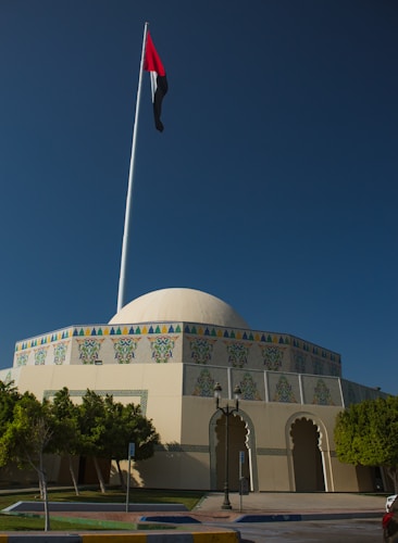A large building with a white dome and ornate decorative tiles. The facade features colorful geometric patterns and traditional archways. A tall flagpole stands in front, displaying a red, green, and black flag. Surrounding the building, there are green trees and a neatly maintained lawn. The sky is clear and blue, providing a striking backdrop.